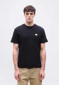 Νεαρός άνδρας με σγουρά μαλλιά, φοράει μαύρο κοντομάνικο μπλουζάκι με υφή και μπεζ παντελόνι, στέκεται μπροστά σε απλό ανοιχτό φόντο.