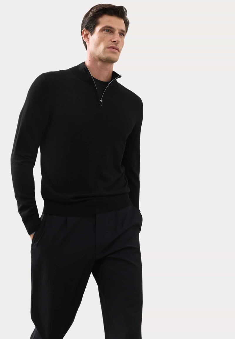 Černý svetr na zip s měkkou pleteninou, dlouhými rukávy a přiléhavým lemem. Nosený s tmavými kalhotami, které zvýrazňují elegantní siluetu.