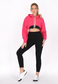 Lyserød cropped hoodie med en sort kant, der har snoretræk. Sammen med højhalset sorte leggings og hvide sneakers med sorte detaljer.