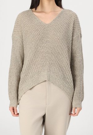 Femme portant un pull en maille beige texturé col en V avec manches longues et ourlet asymétrique, associé à un pantalon beige clair.