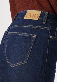 Tumši zili džinsi ar redzamiem šuvēm un gaiši brūnu ādas etiķeti ar uzrakstu "VMD". Iespējas taisnas kabatas un eleganta piegriezuma.