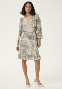 Robe à motifs floraux dans des tons beiges clairs et gris, avec un col en V, des manches longues, un ourlet volanté, et une ceinture en tissu nouée à la taille.