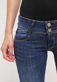 Denim jeans in dark blue with a fitted design. Features include two buttons at the waist, front pocket, and subtle fading on the fabric.