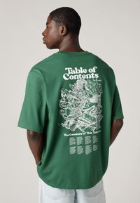 T-shirt verde oversize com texto gráfico branco e ilustrações nas costas, com corte descontraído e mangas curtas.