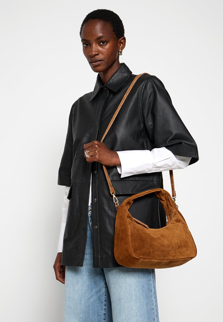 Braune Wildleder-Handtasche mit geschwungener Form, kurzem Tragegurt und strukturiertem Oberflächen, kombiniert mit einem schwarzen Ledershirt und blauen Jeans.
