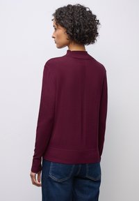 Pull bourgogne avec un col montant, texture côtelée et manches longues. Présente un détail de couture dans le dos, associé à un jean bleu.