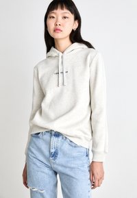 Sweatshirt cinza claro com capuz, apresentando um logótipo frontal. Combinado com jeans rasgados azul claro, usado de forma casual. Textura de tecido suave.