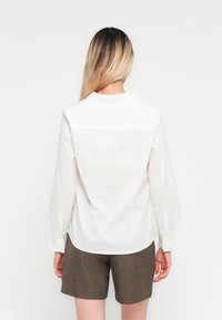 Žena s dlouhými ombre vlasy má na sobě bílou dlouhou blůzu s dlouhým rukávem a hnědé šortky, stojí zády k fotoaparátu na bílém pozadí.