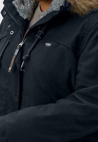 Svart jacka med huva av syntetpäls, med dragkedja, snörning och en tygmärke på bröstet. Mjuk textur och vadderade ärmar.