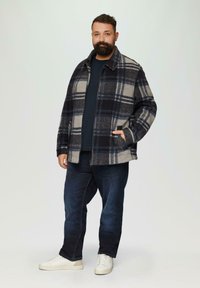 Gecoate blazer in grijs, marineblauw en crème, met een ritssluiting aan de voorkant, twee zijzakken en een kraag, gedragen over een marineblauw shirt en donkere jeans.