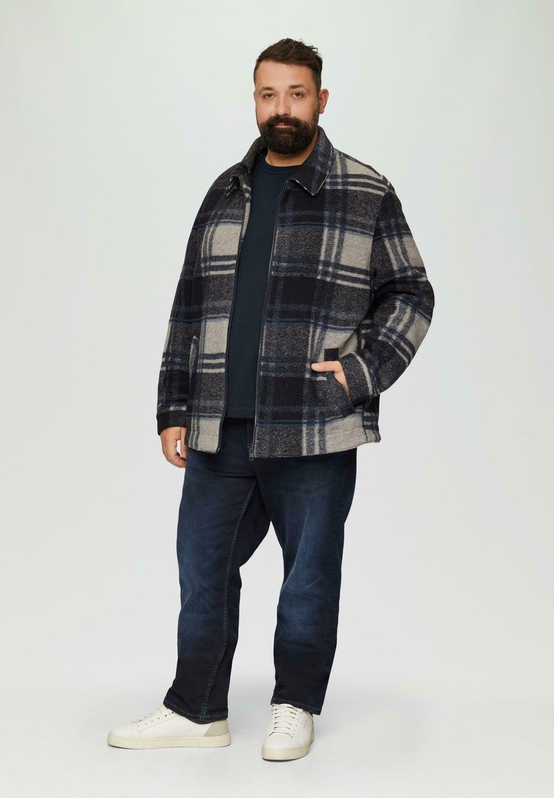 Gecoate blazer in grijs, marineblauw en crème, met een ritssluiting aan de voorkant, twee zijzakken en een kraag, gedragen over een marineblauw shirt en donkere jeans.