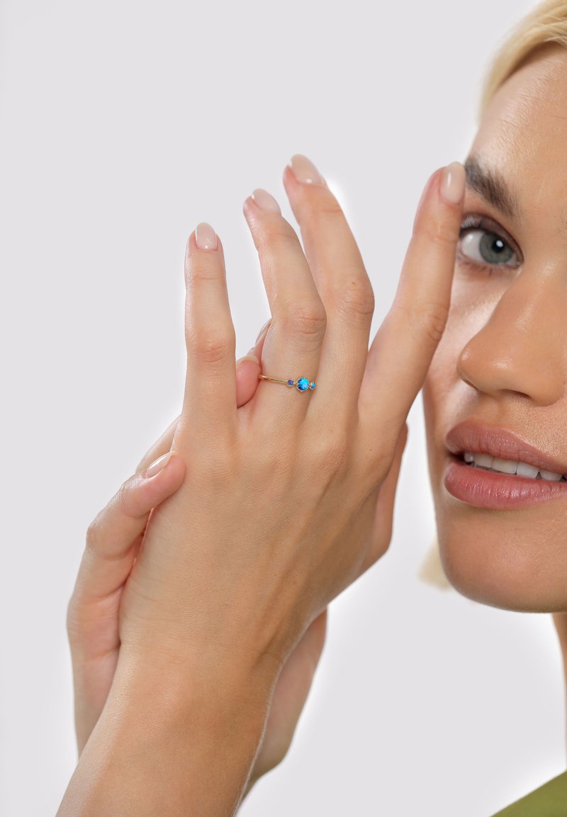 Auksinis žiedas su trimis mėlynais brangakmeniais, smulkia juostele ir blizgiu paviršiumi, rodomas ant rankos su prižiūrėtais nagais.