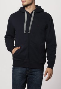 Hombre que lleva una sudadera con cremallera azul marino, con cordones grises y un pequeño logotipo en el pecho, combinada con unos jeans azul oscuro.