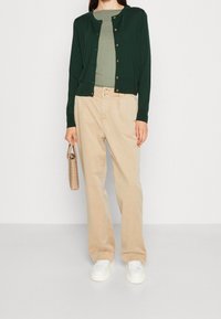 Ciemnozielony kardigan ze złotymi guzikami, nałożony na jasnzielony top, w zestawie z beżowymi spodniami o szerokich nogawkach i białymi trampkami, trzymający beżową torbę.