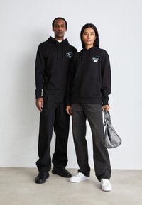 Sudaderas negras con bolsillos frontales y cordones, combinadas con jeans sueltos en negro y gris. Una figura sostiene un bolso gris.