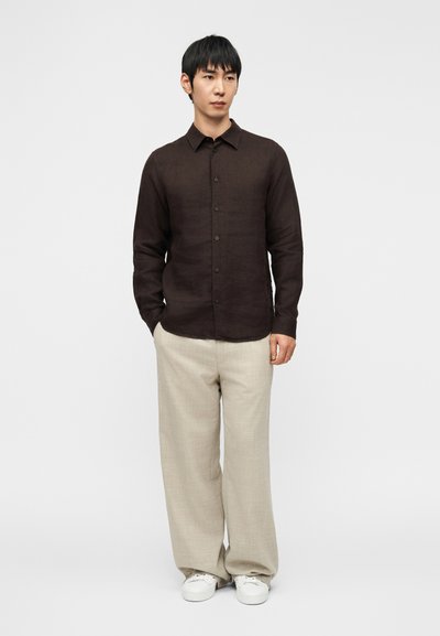 Καφέ πουκάμισο από λινό με κουμπιά και γιακά, σε συνδυασμό με ανοιχτό μπεζ παντελόνι φαρδύ και άσπρα αθλητικά παπούτσια. Απλός σχεδιασμός, λείο υλικό.