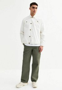 Witte button-up jasje met voorzakken, gecombineerd met olijfgroene cargo broek. Model draagt crèmekleurige sneakers. Eenvoudig, functioneel ontwerp.