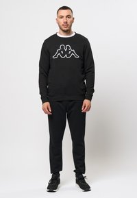 Zwarte sweatshirt met wit Kappa-logo, voorzien van een ronde halslijn en geribde manchetten. Gecombineerd met zwarte sweatpants en zwarte sportschoenen.