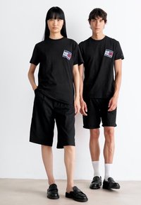 To modeller står side om side iført svarte logo t-skjorter, svarte shorts og svarte slip-on sko foran en enkel hvit bakgrunn.