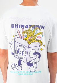 Weißes T-Shirt mit einem Cartoon-Motiv einer Takeout-Box mit Nudeln, Essstäbchen und Garnelen. Der Text lautet "NYC Noodles CHINATOWN."
