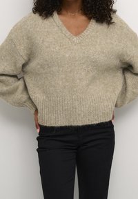 Světlý béžový pletený svetr s výstřihem do V, žebrovanými manžetami a lemem. Nošený přes černé kalhoty, s měkkým a texturovaným povrchem.