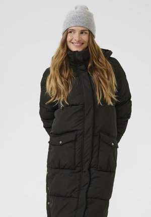 Donna sorridente, che indossa un lungo cappotto imbottito nero e un berretto lavorato a maglia grigio chiaro, in piedi con le mani dietro la schiena su uno sfondo semplice.