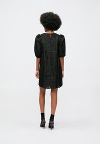 Ženska z afrom, oblečena v teksturirano črno obleko do kolen s napihnjeno rukavico in črnimi petkami, stoji pred enobarvnim ozadjem.