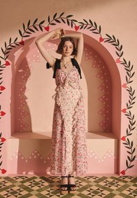 Ung kvinna i en blommig rosa klänning med svarta rosettar på axelbanden och platåsandaler poserar med armarna upplyfta framför en dekorativ rosa båge med hjärtan.