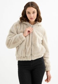 Chaqueta de felpa sintética beige con cremallera y capucha, que cuenta con una cinturilla fruncida y una superficie texturizada. Modelo con jeans negros, presentando un look casual.