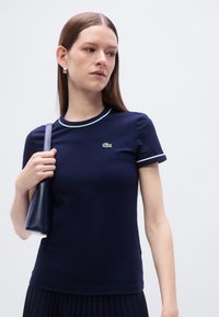 Μπλε ναυτικό κοντομάνικο μπλουζάκι με λευκή διακόσμηση και λογότυπο Lacoste στο στήθος, φτιαγμένο από λείο ύφασμα, σε συνδυασμό με μια μικρή τσάντα.