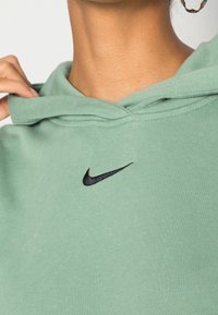 Person som bär en grön huvtröja med en svart Nike-logga, håller huvan vid nacken, med en ringörhänge synlig i ena örat.