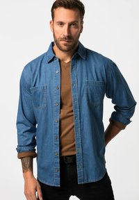 Mediumpuna denim-paita pitkällä hihalla, napit edessä, rintataskut ja käärityt hihat, kerrostettuna ruskean V-päällyspaidan päällä.