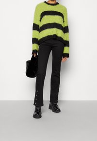 Fluffy groene en zwart gestreepte trui, zwarte strakke jeans met drukknoopdetails aan de zoom, en zwarte chunky schoenen, gecombineerd met een zwarte tas.