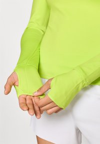 Φωτεινό πράσινο μπλούζα μακριά μανίκια με τ�ρύπες για τους αντίχειρες, λεία υφή, σχεδίαση που αγκαλιάζει το σώμα και διακριτικές λεπτομέρειες στα μανίκια.