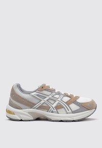 Zapatilla deportiva gris y beige con materiales de malla y ante, suela blanca, detalles reflectantes plateados y cierre de cordones.