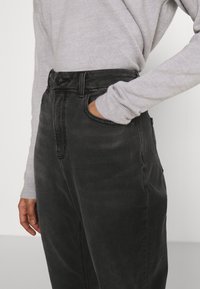 Jeans de denim negro de cintura alta con un bolsillo frontal, cierre de cremallera y trabillas para cinturón, combinados con un suéter de manga larga gris claro.