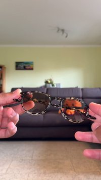 Gafas de sol redondas con un patrón de carey negro y gris, lentes oscuros y delgadas varillas de metal, sostenidas en las manos de una persona.