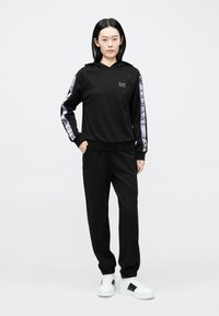 Czarna bluza z kapturem i dopasowane spodnie wykonane z miękkiego materiału. Bluza posiada kaptur, unikalne, wzorzyste rękawy oraz białe sneakersy z czarnymi akcentami.