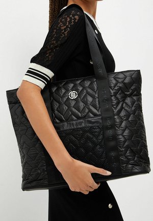 Sac fourre-tout noir matelassé avec des sections lisses et texturées, accent logo argenté et bretelles noires robustes avec marquage en relief.
