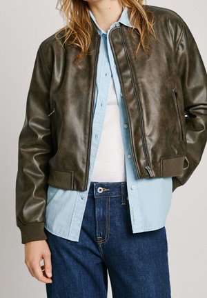 Persoon die een donkergroene leren jas draagt over een lichtblauw overhemd met knopen, een wit topje en blauwe spijkerbroek, staand met één hand langs het zij.