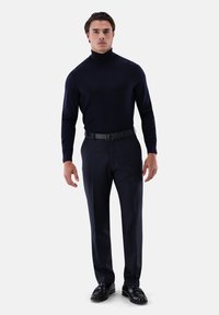 Marineblauer Rollkragenpullover aus weichem Stoff, kombiniert mit maßgeschneiderten dunklen Hosen und schwarzen Loafers. Modell steht vor einem weißen Hintergrund.