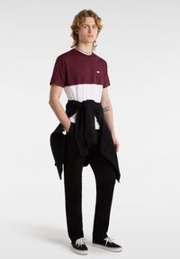 Bordeaux- und weißes Color-Block-T-Shirt mit kleinem Logo, kombiniert mit schwarzen Jogginghosen und Schuhen, und einer schwarzen Kapuzenjacke, die um die Taille gebunden ist.