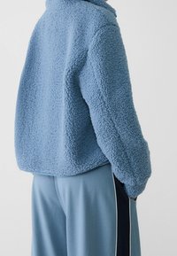 Chlupatá modrá fleecová bunda s límcem a oversize střihem, s texturovaným povrchem, dlouhými rukávy a kulatým lemem.