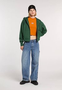 Grøn hættetrøje over en orange kort top, kombineret med løse, lyseblå jeans og sorte slip-on sko. Simpelt design, afslappet pasform.