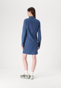 Vestido de ganga em azul escuro, com mangas longas, cintura ajustada, punhos com botões e comprimento até a meia coxa. Usado com meias claras e ténis brancos com detalhes.