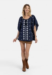 Tunic blu navy con ricami floreali bianchi, ampie maniche a pipistrello e scollatura con cordoncino, abbinata a una mini gonna in suede marrone e stivali.