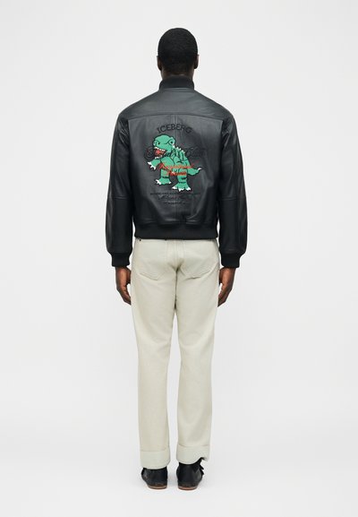 Άνδρας που φοράει μαύρο δερμάτινο μπουφάν με πολύχρωμο καρτούν δεινόσαυρο και κείμενο στην πλάτη, συνδυασμένο με ανοιχτό μπεζ παντελόνι και μαύρα παπούτσια.