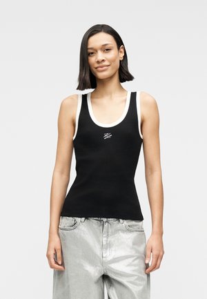 Débardeur noir côtelé avec bordure blanche, présentant un col boule et un logo brodé au centre. Porté avec un pantalon argenté métallique.