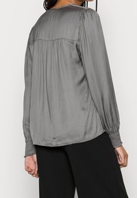 Blusa satin grigia con maniche lunghe, dettaglio arricciato lungo il collo e sul retro, e polsini smock. Indossata con pantaloni neri a gamba larga.
