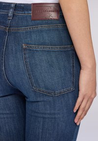 Jeans de mezclilla de color azul oscuro con un bolsillo trasero, que presentan un contraste de costuras naranjas y una etiqueta de cuero marrón en la cintura.